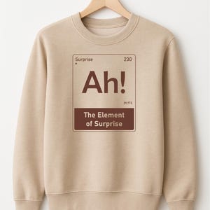 May include: A beige sweatshirt with a brown graphic that resembles a periodic table element. The graphic reads "Ah! The Element of Surprise". The sweatshirt is hanging on a wooden hanger.