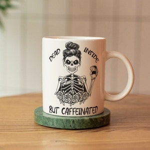 May include: White ceramic coffee mug with a black and white illustration of a skeleton with a messy bun holding a coffee cup. The text on the mug reads "Dead Inside But Caffeinated".