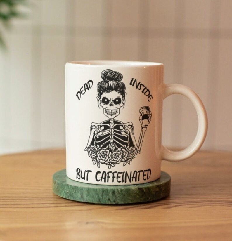 May include: White ceramic coffee mug with a black and white illustration of a skeleton with a messy bun holding a coffee cup. The text on the mug reads "Dead Inside But Caffeinated".