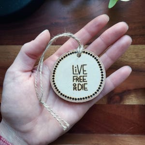 May include: A round, wooden ornament with the words "LIVE FREE OR DIE" burned into the surface. The ornament has a dotted border and a twine loop for hanging. The wood has a natural, light color.