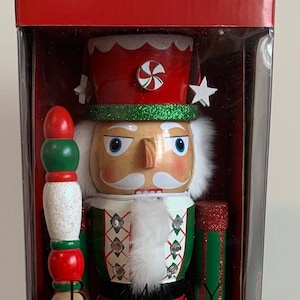 May include: A red, green and white wooden nutcracker with a candy cane hat and a white beard. The nutcracker is holding a candy cane stick and is standing in a clear plastic box with a red lid.