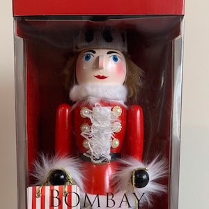 May include: A red and white striped shopping bag with the text "Bombay" hanging from the arm of a wooden nutcracker. The nutcracker is wearing a red and white uniform with a white fur trim and a grey and white striped stocking. The nutcracker is holding a red and white drum with a black rim. The nutcracker is standing on a black and white chequered base.
