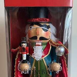 May include: A wooden nutcracker in a red and blue costume with gold trim and a black eyepatch. The nutcracker is holding a silver ball on a stick in one hand and a smaller nutcracker in the other. The nutcracker is in a clear plastic box with a red lid. The box has the word "Bombay" printed on it.