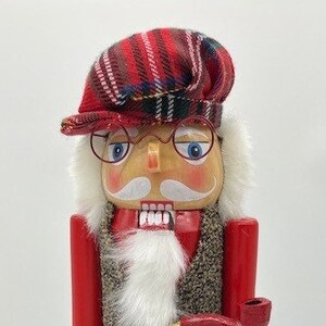 May include: A wooden nutcracker figure wearing a red and plaid hat, spectacles, and a tweed waistcoat. The figure has a white beard, red arms, black trousers, and red boots with green and red details. It holds a red pipe.