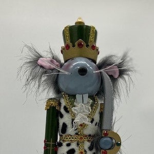 May include: A grey mouse nutcracker with a green and gold crown, holding a silver sword. The nutcracker is wearing a white and black spotted coat with gold trim and red trousers. The nutcracker is standing on a red and gold base.