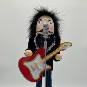 May include: A wooden nutcracker dressed as a rock star with long black hair, a black leather jacket, and blue jeans. He is holding a red and white electric guitar and a microphone. He is standing on a stage with a gold base and musical notes.