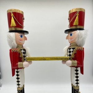 May include: Two red and gold nutcracker figurines holding a gold tray. The nutcrackers are wearing black hats with gold trim and white fur. They are standing on a gold and black base.
