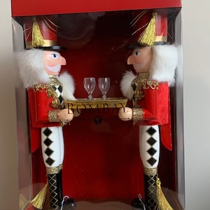 May include: Two red and gold nutcracker figurines holding a tray with two wine glasses. The nutcrackers are wearing white and black diamond patterned pants and have white fur trim. The box they are in is red and says "Bombay" on the front.