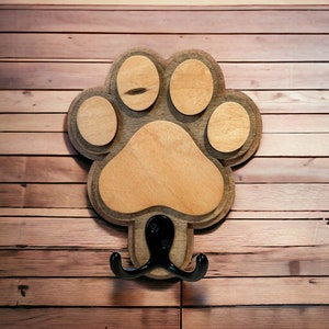 May include: Wooden dog paw print hook with a black metal hook. The hook is attached to the paw print and is shaped like a double hook.