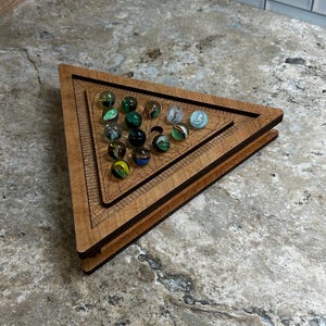 May include: A wooden triangular marble game board with multiple marbles in various colors. The board is made of layered wood and has a detailed design. The marbles are clear, green, blue, and black.