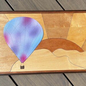 Può includere: Un'opera d'arte in legno che raffigura una mongolfiera con un design viola e blu su uno sfondo di tramonto. La mongolfiera è in primo piano e il tramonto è sullo sfondo. Il pezzo è incorniciato in una cornice di legno scuro.