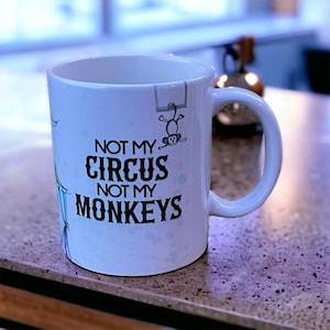 Puede incluir: Taza de café de cerámica blanca con un gráfico en blanco y negro que dice "No es mi circo, no son mis monos". Un pequeño mono está colgado de un trapecio sobre el texto.