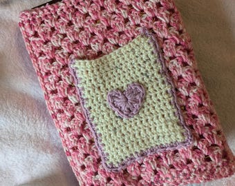 Puede incluir: Una cubierta de libro de crochet rosa y blanco con un pequeño bolsillo beige con un corazón morado. La cubierta está sobre una superficie blanca y peluda.