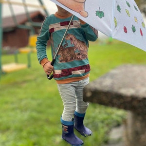 May include: A child wearing a blue and orange long-sleeved shirt with a dinosaur graphic. The shirt has a red and white trim. The child is holding a blue umbrella with a bear and leaves printed on it. The child is wearing blue rain boots.