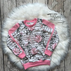May include: A pink and gray striped sweatshirt with a floral print and keyhole design. The sweatshirt has pink sleeves and a pink waistband. The text "Tüpfel fuchs" and "Freestyle" are visible on the image.