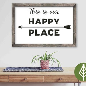 May include: A framed sign with a white background and black text that reads "This is our HAPPY PLACE" with a black arrow pointing to the right. The sign is hanging on a white wall above a wooden table with a green plant in a red and white pot.