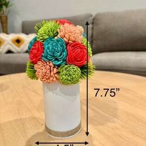 May include: A colorful floral arrangement in a white vase. The bouquet features red, teal, green, and peach-colored flowers. The vase is 4.5 inches wide and 7.75 inches tall, with a jute rope accent around the base. The arrangement sits on a wooden table.