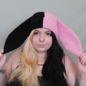 Pode incluir: Um gorro de crochê bicolor com longas orelhas de coelho caídas. O gorro é dividido verticalmente ao centro, com um lado em preto sólido e o outro em rosa claro sólido, confeccionado com uma textura canelada em toda a peça.