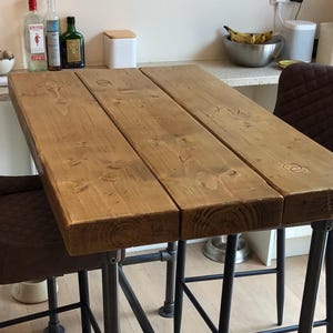 May include: A rustic wooden bar table with a natural wood finish and black metal legs. The table is set up with two brown leather bar stools.
