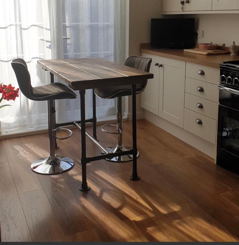 May include: A rustic kitchen island with a dark wood tabletop and black metal pipe legs. Two gray upholstered bar stools with chrome bases are positioned at the island. The island is in front of a window with sheer curtains.