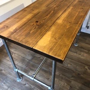 May include: A rectangular wooden table with a warm brown finish. The tabletop is constructed from three planks. The table is supported by industrial-style metal pipe legs and crossbars, creating a sturdy and stylish design.
