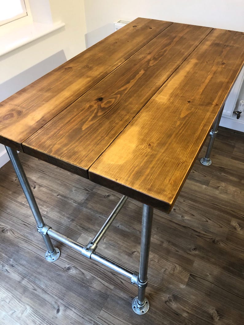 May include: A rectangular wooden table with a warm brown finish. The tabletop is constructed from three planks. The table is supported by industrial-style metal pipe legs and crossbars, creating a sturdy and stylish design.