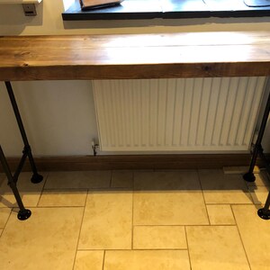 May include: A wooden countertop with black metal pipe legs. The countertop is brown and the legs are black. The countertop is mounted to a wall and is about 3 feet long.