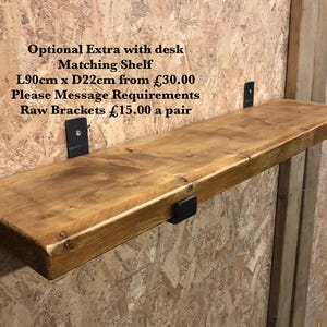 May include: A wooden shelf, approximately 35 inches long and 8.7 inches deep, mounted on black metal brackets. The shelf has a natural wood grain finish and is attached to a light brown wall. Text on the wall reads "Optional Extra with desk Matching Shelf".