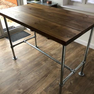 May include: A rectangular wooden table with a dark brown finish. The table is supported by galvanized steel pipes and fittings. The table has a rustic, industrial design and is suitable for various uses.