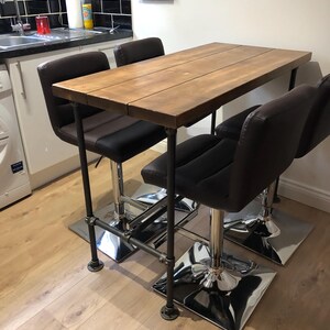 May include: A rustic bar table with a wooden top and black metal pipe legs. Four black and brown leather bar stools with chrome bases are positioned around the table. The table is approximately 48 inches long.