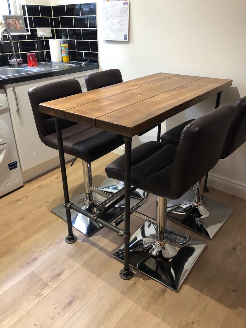May include: A rustic bar table with a wooden top and black metal pipe legs. Four black and brown leather bar stools with chrome bases are positioned around the table. The table is approximately 48 inches long.