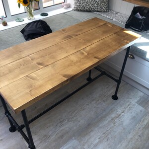 May include: A rectangular wooden table with a light brown finish. The tabletop is constructed from several planks. The table has black metal pipe legs and supports. The table is approximately 60 inches long and 30 inches wide.