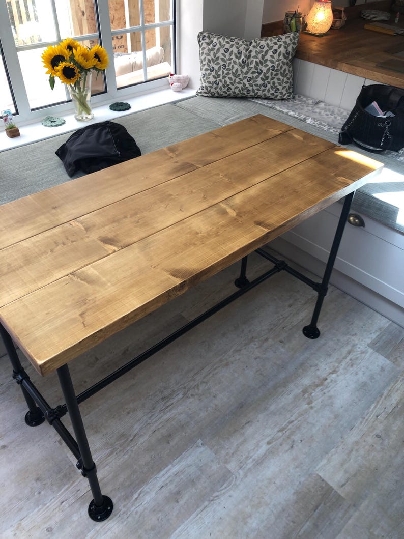 May include: A rectangular wooden table with a light brown finish. The tabletop is constructed from several planks. The table has black metal pipe legs and supports. The table is approximately 60 inches long and 30 inches wide.