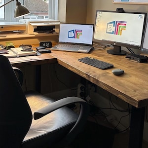 May include: A home office setup featuring an L-shaped wooden desk with a black office chair, a laptop, two computer monitors, a keyboard, a mouse, and a desk lamp. The screens display a logo with the letters CIF and colourful horizontal bars.