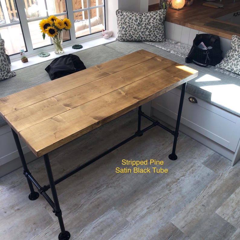 May include: A rectangular wooden table with a light brown finish. The table is supported by black metal pipe legs. The text "Stripped Pine Satin Black Tube" is visible on the image.