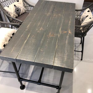 May include: A dark gray wooden table with a distressed finish and black metal pipe legs. The table is set with two chairs with woven backs and white and gray patterned cushions.
