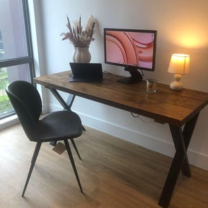 May include: A rustic wooden desk with black metal legs, a laptop, a monitor, a vase with dried flowers, and a lamp. The desk is in a room with light wood floors.