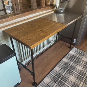 May include: A rustic kitchen island with a wooden countertop and a stainless steel extension. The island has a black metal pipe frame and sits on a wooden floor. A light blue trash can is visible on the left.