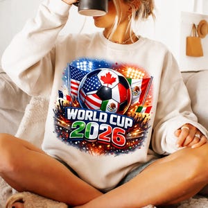 May include: Cream-colored sweatshirt featuring a soccer ball with flags of the USA, Canada, and Mexico. The design includes the text "WORLD CUP 2026" in green and red, with a fireworks-like background.