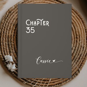 May include: A dark gray journal with the words "CHAPTER 35" in white script. The name "Cassie" is written in cursive below, with a small heart. The journal is resting on a woven, circular placemat.