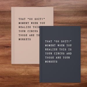 May include: Two notebooks, one beige and one dark gray, with the text "THAT 'OH SHIT!' MOMENT WHEN YOU REALIZE THIS IS YOUR CIRCUS AND THOSE ARE YOUR MONKEYS" printed on the covers. The notebooks are on a wooden surface.