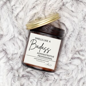 Puede incluir: Un tarro de vidrio marrón con una vela y una tapa dorada. La etiqueta dice "Smells like a Badass Administrative Professional" y "Hand poured with love".
