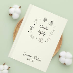 Puede incluir: Libro de tapa dura color crema con el texto "Chapter Eighty" rodeado de ilustraciones lineales de iconos de celebración. La portada del libro también incluye el nombre "Cameron Packer" y el año "EST 1946".