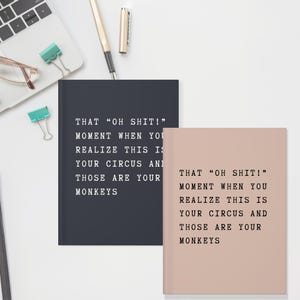 May include: Two notebooks, one navy blue and one blush pink, with the text "THAT 'OH SHIT!' MOMENT WHEN YOU REALIZE THIS IS YOUR CIRCUS AND THOSE ARE YOUR MONKEYS" printed in black. A laptop, pen, and paper clips are in the background.