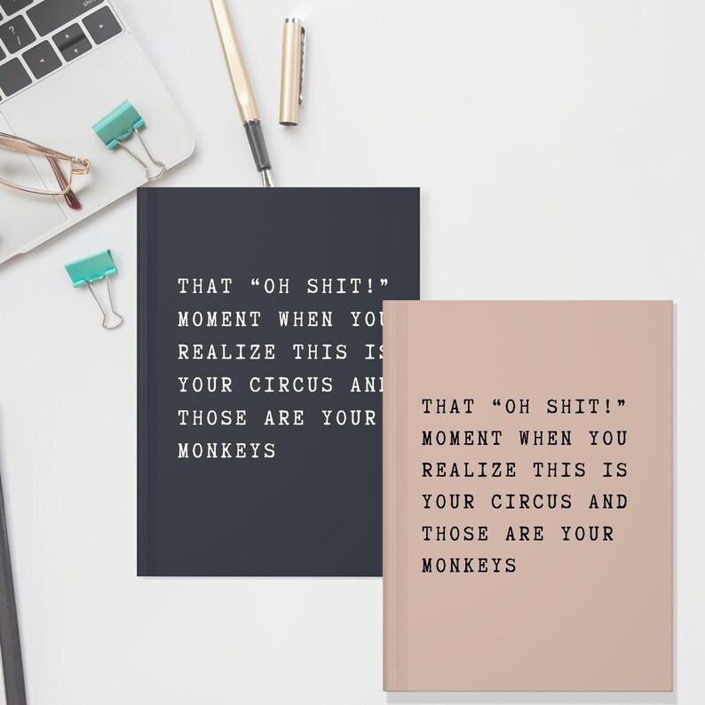 May include: Two notebooks, one navy blue and one blush pink, with the text "THAT 'OH SHIT!' MOMENT WHEN YOU REALIZE THIS IS YOUR CIRCUS AND THOSE ARE YOUR MONKEYS" printed in black. A laptop, pen, and paper clips are in the background.