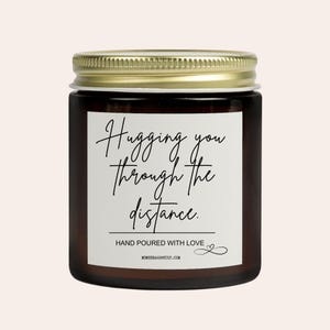 May include: A brown glass jar candle with a gold lid. The label reads "Hugging you through the distance." Below the text is "HAND POURED WITH LOVE" and the website address. The candle is a home decor item.