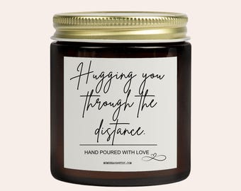 May include: A brown glass jar candle with a gold lid. The label reads "Hugging you through the distance." Below the text is "HAND POURED WITH LOVE" and the website address. The candle is a home decor item.