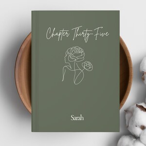 May include: A hardcover book with a sage green cover. The cover features the text "Chapter Thirty-Five" in cursive, a line drawing of a woman's profile with flowers, and the name "Sarah" below. The book is resting in a wooden bowl.