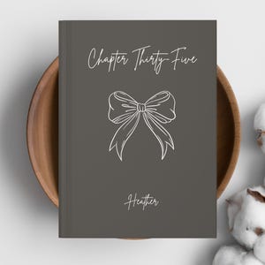 May include: A gray book with the text "Chapter Thirty-Five" and the name "Heather" in white script. A white bow illustration is centered on the cover. The book is in a wooden bowl.