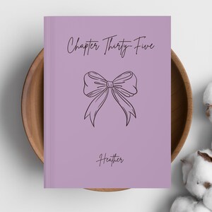 May include: A lavender-colored book with the text "Chapter Thirty-Five" and the name "Heather" in a handwritten font. A line drawing of a bow is in the center of the cover. The book is in a wooden bowl.